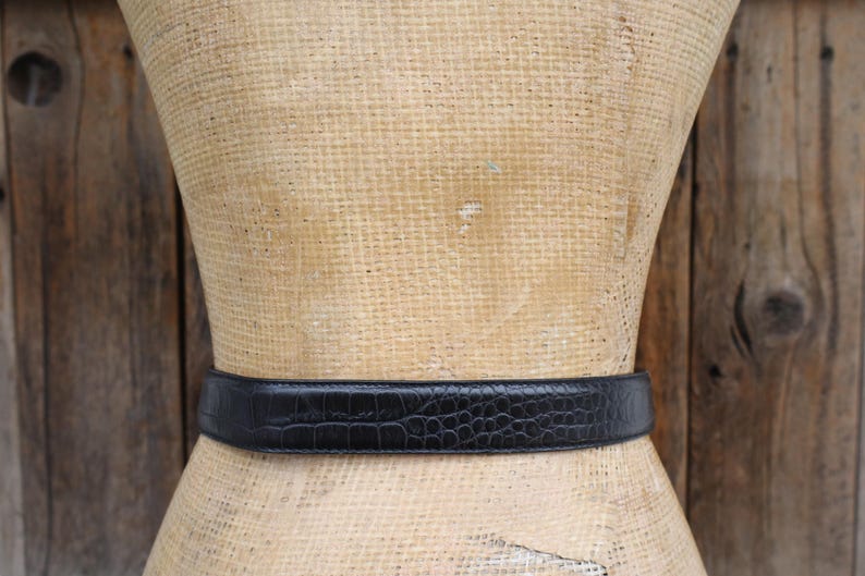 May include: A black belt with a crocodile-like texture is wrapped around a mannequin. The belt is made of a dark material and has a rectangular buckle. The background is a wooden surface.