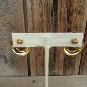 K&ouml;nnte beinhalten: Ein Paar goldfarbene Ohrringe mit einem runden Design. Die Ohrringe werden auf einem wei&szlig;en Stoffst&auml;nder pr&auml;sentiert.