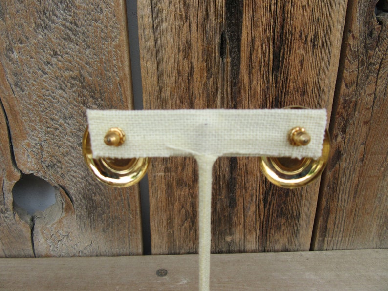 K&ouml;nnte beinhalten: Ein Paar goldfarbene Ohrringe mit einem runden Design. Die Ohrringe werden auf einem wei&szlig;en Stoffst&auml;nder pr&auml;sentiert.