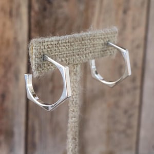 May include: A pair of silver-colored, geometric hoop earrings. Each earring has a hexagonal shape with a flat, polished surface. The earrings are displayed on a tan fabric earring stand against a blurred wooden background.