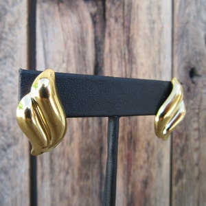 Puede incluir: Un par de pendientes de tono dorado con un diseño de hoja. Los pendientes se muestran en un soporte negro.