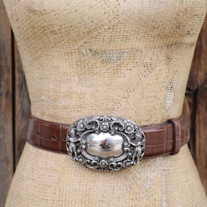 May include: A brown leather belt with a silver oval buckle. The buckle has an ornate design with two figures on either side of a central silver plate. The belt is displayed on a mannequin.