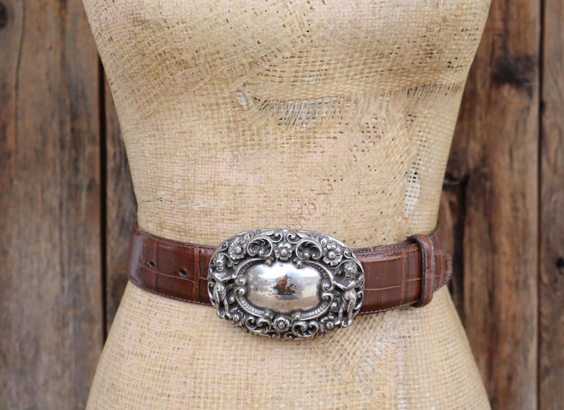 May include: A brown leather belt with a silver oval buckle. The buckle has an ornate design with two figures on either side of a central silver plate. The belt is displayed on a mannequin.
