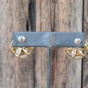 K&ouml;nnte beinhalten: Ein Paar goldene Ohrringe mit einem rosafarbenen Opal-Stein, der in einem geometrischen Design gefasst ist. Die Ohrringe werden auf einem schwarzen St&auml;nder pr&auml;sentiert.