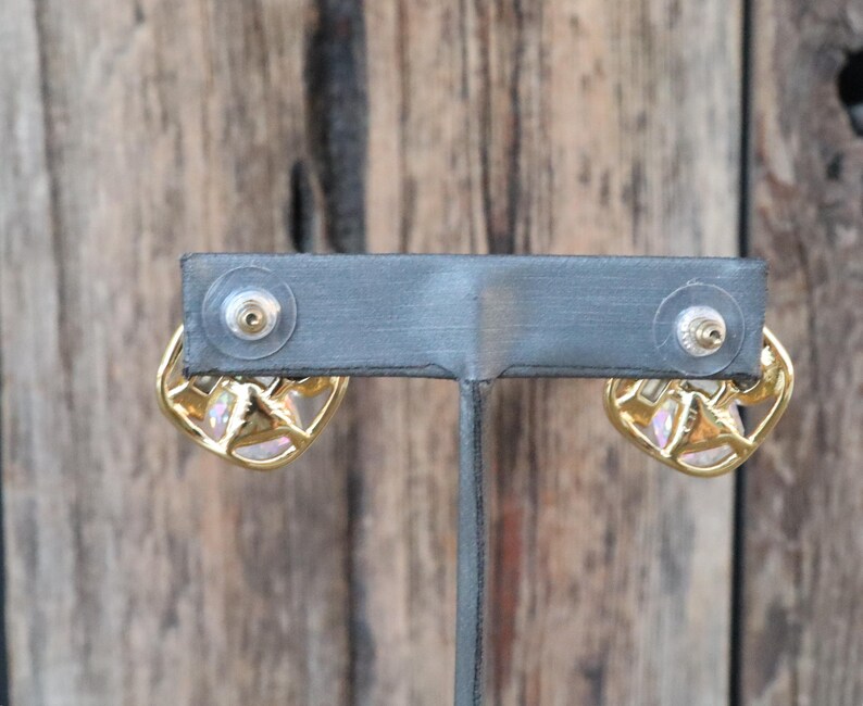 K&ouml;nnte beinhalten: Ein Paar goldene Ohrringe mit einem rosafarbenen Opal-Stein, der in einem geometrischen Design gefasst ist. Die Ohrringe werden auf einem schwarzen St&auml;nder pr&auml;sentiert.