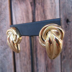 May include: A pair of gold-tone knot-shaped earrings. The earrings are on a black display stand.