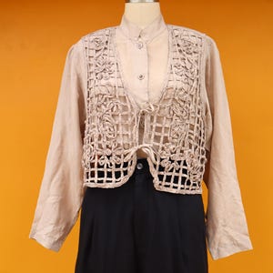 May include: A beige, open-weave vest with floral detailing over a long-sleeved beige shirt and black pants. The vest has a grid-like pattern with floral accents. The outfit is displayed on a mannequin against an orange background.