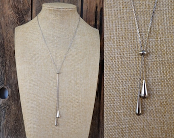 Peut inclure: Un collier argenté avec un design en forme de Y. Le collier présente une fine chaîne menant à un petit accent rond, puis se divise en une chaîne plus longue avec deux pendentifs en forme de goutte. Le bijou est exposé sur un mannequin.