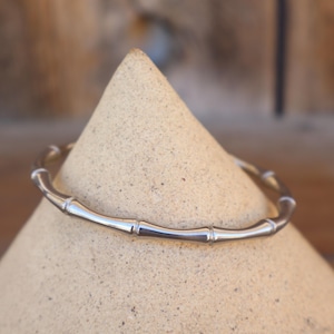 May include: A silver-colored bangle bracelet with a bamboo design. The bracelet is displayed on a beige, cone-shaped object. The bamboo-style bracelet has several segments and is made of a shiny metal.