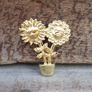 May include: A gold-colored metal brooch in the shape of two sunflowers in a pot. The sunflowers have detailed petals and are connected by stems and leaves. The pot is also gold and has a simple design.