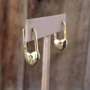 May include: A pair of gold-colored earrings shaped like padlocks with heart-shaped bases. The earrings are displayed on a beige earring stand against a blurred wooden background. Each padlock has a keyhole detail.