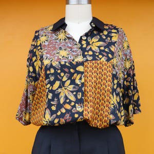 May include: A collared blouse with a patchwork design featuring floral and geometric patterns in navy, brown, and yellow. The blouse has a black collar and is paired with black trousers.