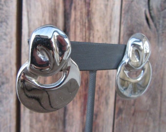 May include: A pair of silver tone earrings with a unique abstract design. The earrings are on a black display stand.