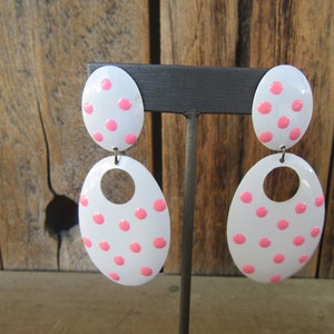 May include: A pair of white oval earrings with pink polka dots. The earrings are dangle style with a smaller oval on top of a larger oval.