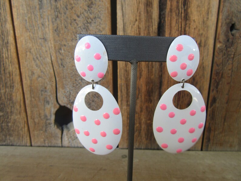 May include: A pair of white oval earrings with pink polka dots. The earrings are dangle style with a smaller oval on top of a larger oval.