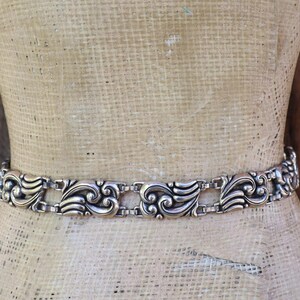 May include: Silver-toned metal belt with an ornate, repeating scroll design. The belt is displayed on a mannequin, showcasing its intricate details and vintage style. The belt's design features a series of stylized, symmetrical floral and leaf motifs.