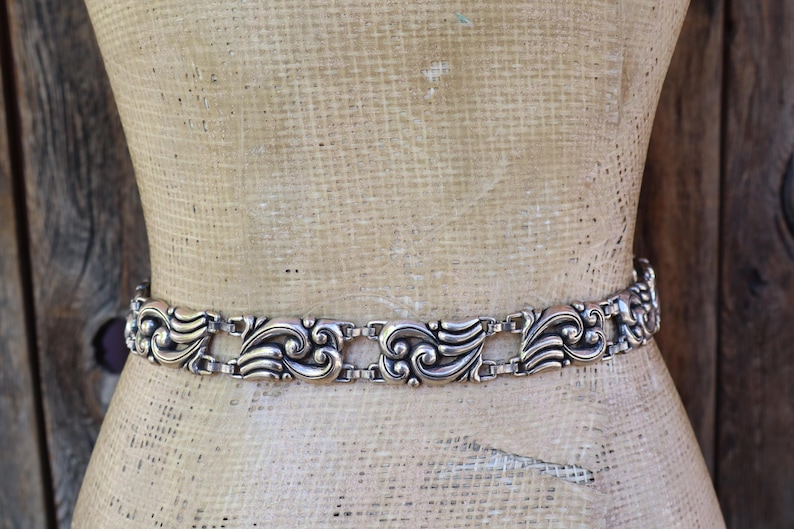 May include: Silver-toned metal belt with an ornate, repeating scroll design. The belt is displayed on a mannequin, showcasing its intricate details and vintage style. The belt's design features a series of stylized, symmetrical floral and leaf motifs.