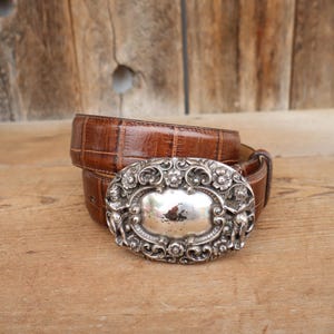 May include: A brown leather belt with a silver-colored, ornate buckle. The buckle has a central oval design surrounded by floral and scrollwork details. The belt is made of textured leather.