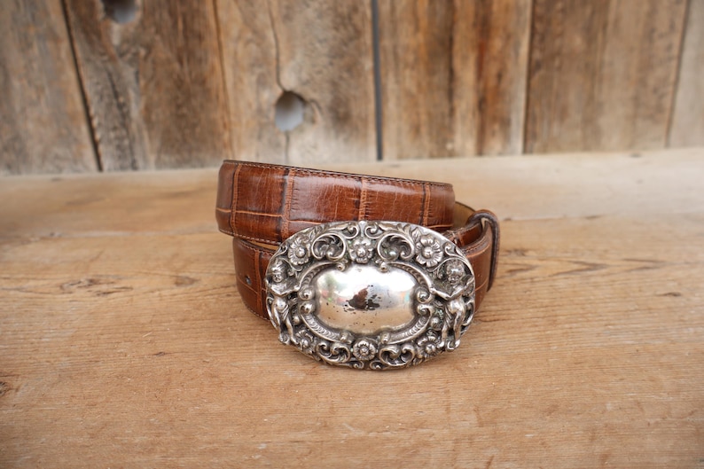 May include: A brown leather belt with a silver-colored, ornate buckle. The buckle has a central oval design surrounded by floral and scrollwork details. The belt is made of textured leather.
