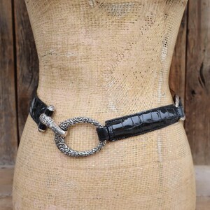 May include: Black belt with a silver-toned buckle. The belt has a glossy, crocodile-like texture. The buckle is a large, ornate oval shape. The belt is displayed on a mannequin.