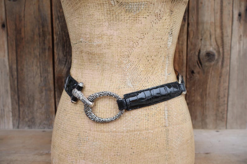 May include: Black belt with a silver-toned buckle. The belt has a glossy, crocodile-like texture. The buckle is a large, ornate oval shape. The belt is displayed on a mannequin.