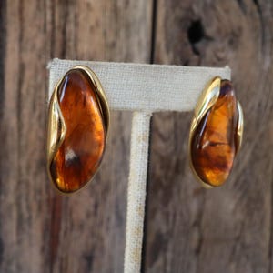 Può includere: Un paio di orecchini vintage con una pietra ovale color ambra, incastonata in una montatura in metallo dorato. Gli orecchini sono esposti su un supporto in tessuto neutro, su uno sfondo sfocato in legno. Finitura lucida.