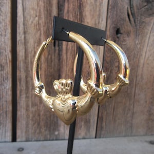 May include: Gold-toned hoop earrings featuring a Claddagh design. The earrings have a heart at the center, clasped by two hands and topped with a crown. The hoops are displayed against a wooden background.