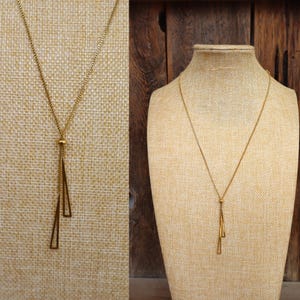 May include: A gold-colored necklace with a delicate chain and a pendant featuring two elongated, open geometric shapes. The necklace is displayed on a neutral-toned mannequin, showcasing its design and length. The jewelry is simple and elegant.