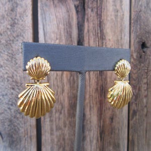 May include: A pair of gold-tone shell-shaped dangle earrings. The earrings are on a black display stand.