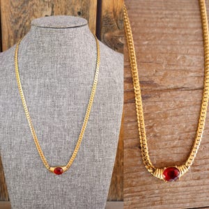 May include: A gold-toned chain necklace featuring a flat, interlocking link design. The necklace is centered with a single, oval-shaped, deep red gemstone set in a gold-toned metal frame that connects the two sides of the chain.