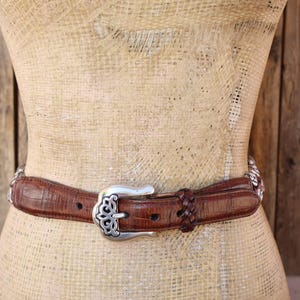 May include: Brown leather belt with a silver buckle and decorative accents. The belt features a textured surface and a braided detail near the buckle. The buckle has an ornate design.