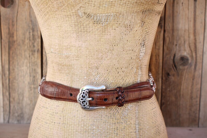 May include: Brown leather belt with a silver buckle and decorative accents. The belt features a textured surface and a braided detail near the buckle. The buckle has an ornate design.