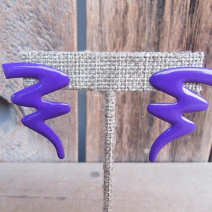 May include: A pair of vibrant purple earrings with a zig-zag design. The earrings are made of a glossy material and are displayed on a neutral-toned stand. These statement earrings are a bold accessory.