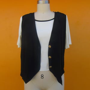 Peut inclure: Un gilet noir sans manches avec des boutons dor&eacute;s. Le gilet a une ourlet pointu et est port&eacute; sur un t-shirt blanc.