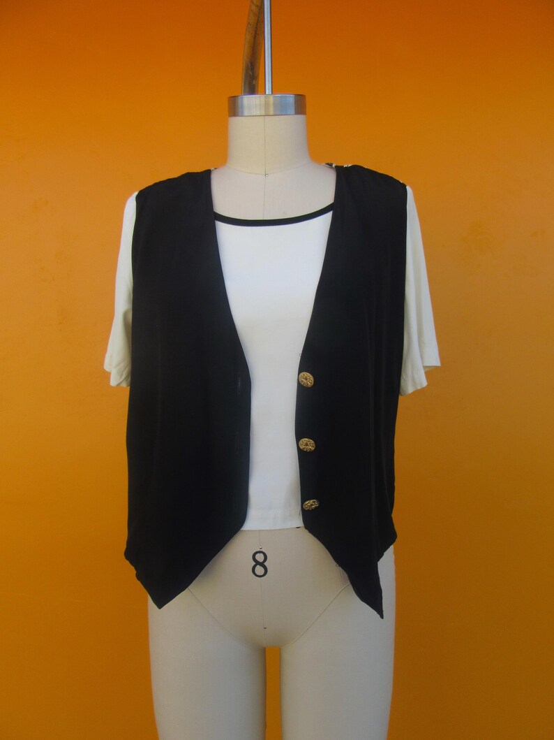 Peut inclure: Un gilet noir sans manches avec des boutons dor&eacute;s. Le gilet a une ourlet pointu et est port&eacute; sur un t-shirt blanc.