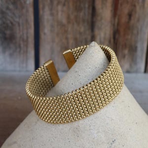 Può includere: Un braccialetto a rete color oro con chiusura rettangolare. Il braccialetto è esposto su un oggetto chiaro a forma di cono. Lo sfondo è una superficie in legno.