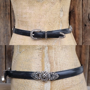 May include: Two black leather belts with silver-toned buckles and decorative accents. The top belt features a classic buckle, while the bottom belt has a unique center piece with heart-shaped details. Both belts are displayed on a mannequin.