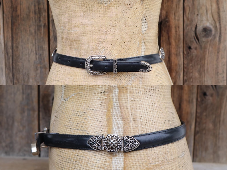 May include: Two black leather belts with silver-toned buckles and decorative accents. The top belt features a classic buckle, while the bottom belt has a unique center piece with heart-shaped details. Both belts are displayed on a mannequin.