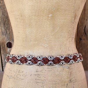 May include: A brown leather belt with a woven design and silver-colored metal accents. The belt is displayed on a mannequin, showcasing its intricate details and craftsmanship. The belt is a fashion accessory.