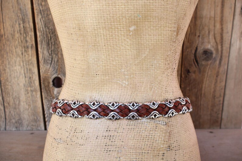May include: A brown leather belt with a woven design and silver-colored metal accents. The belt is displayed on a mannequin, showcasing its intricate details and craftsmanship. The belt is a fashion accessory.