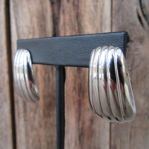 May include: A pair of silver hoop earrings with a ridged design. The earrings are displayed on a black stand.