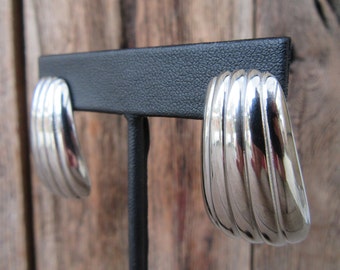 May include: A pair of silver hoop earrings with a ridged design. The earrings are displayed on a black stand.