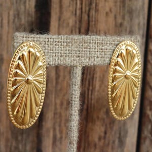 May include: A pair of gold oval-shaped earrings with a sunburst design. The earrings are on a burlap background.