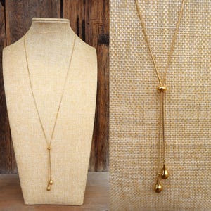 May include: A gold-colored necklace with a delicate chain and a lariat design. The necklace features a small, round bead and two teardrop-shaped pendants. The jewelry is displayed on a mannequin against a neutral background.