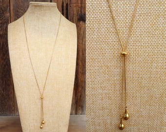 Può includere: Una collana color oro con una catena delicata e un design a lariat. La collana presenta una piccola perla rotonda e due pendenti a forma di goccia. Il gioiello è esposto su un manichino su uno sfondo neutro.