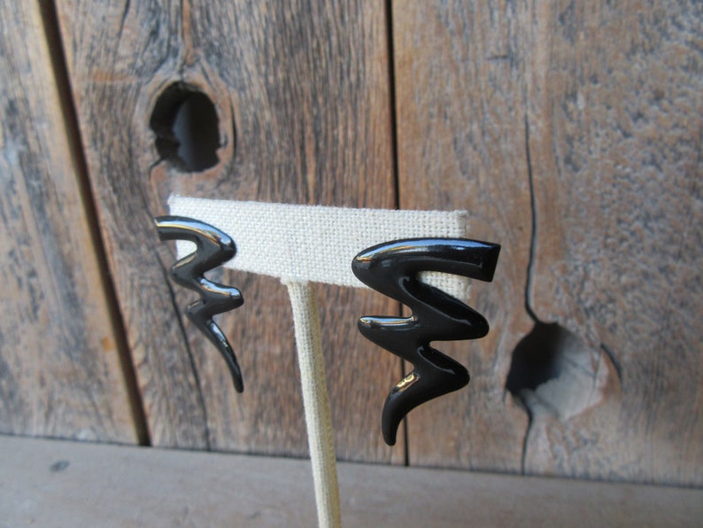 Pode incluir: Um par de brincos pretos de forma geom&eacute;trica abstrata. Os brincos s&atilde;o feitos de um material brilhante e liso e s&atilde;o exibidos em um suporte branco.