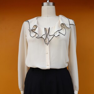May include: Cream-colored blouse with a ruffled collar trimmed in black. The long-sleeved top has a button-down front and is paired with black trousers. The blouse is displayed on a mannequin against an orange background.