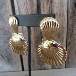 May include: A pair of gold-toned metal earrings featuring a double shell design. Each earring consists of a smaller, vertically oriented shell shape attached to a larger, rounded fan-shaped shell, both with a polished finish and ribbed texture.