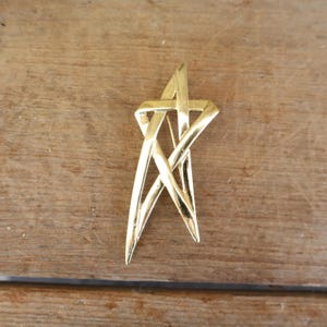 Könnte beinhalten: Goldfarbene sternförmige Brosche mit abstraktem Design. Die Brosche hat sich kreuzende Linien, die ein Sternmuster bilden. Die Brosche besteht aus glänzendem Metall und liegt auf einem Holzgrund.