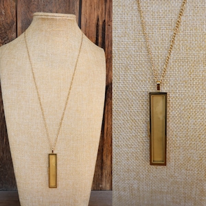 Puede incluir: Un collar dorado con una cadena larga y un colgante rectangular. El colgante tiene un marco dorado y un interior de color claro. El collar se exhibe en un maniquí beige.